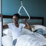 A woman using a SuperPole with a SuperTrapeze to pull herself to a sitting position while laying down in bed.